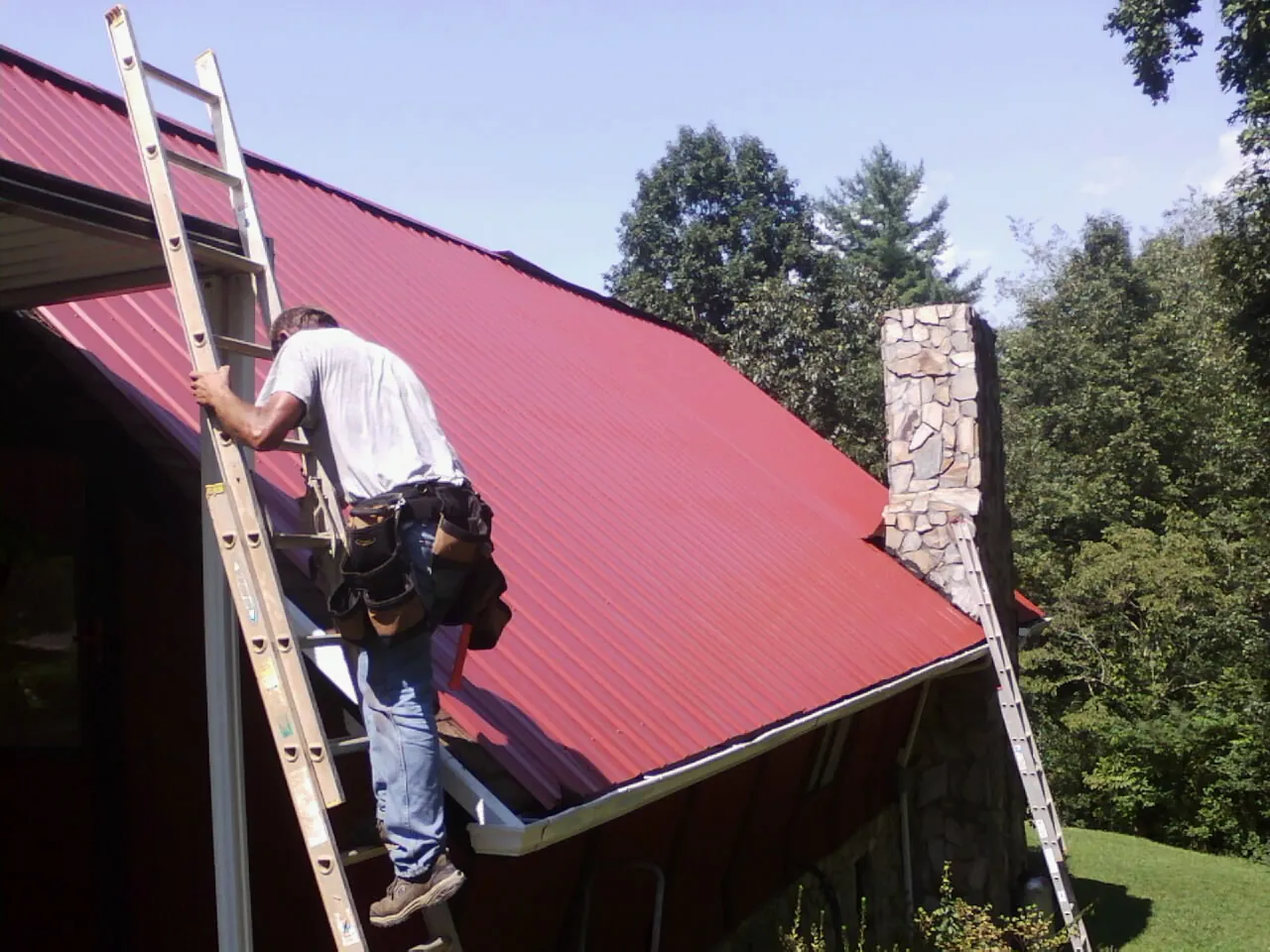Roofer on a ladder beside a red metal roof and stone chimney — Soffit & Fascia Repair by Roofing Brothers in Dansville